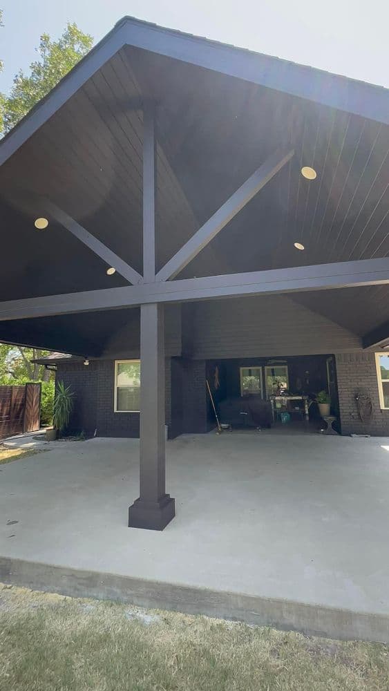 Modern patio with a dark wooden beam structure and concrete floor, surrounded by greenery.