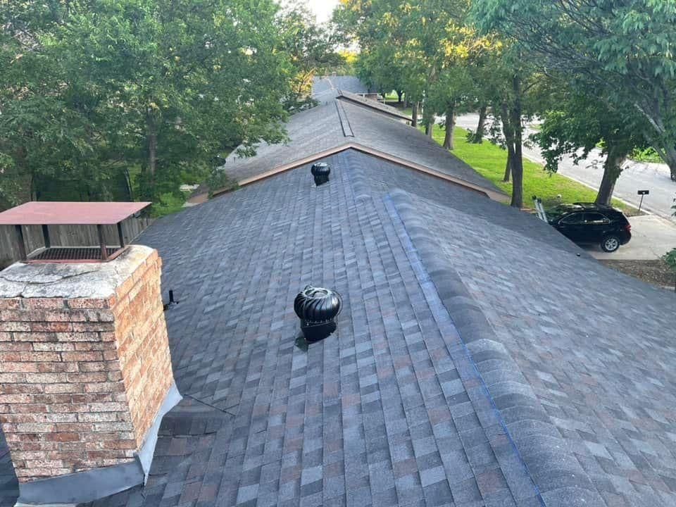 Aerial view of a residential roof with shingles, chimney, and ventilation system among trees.