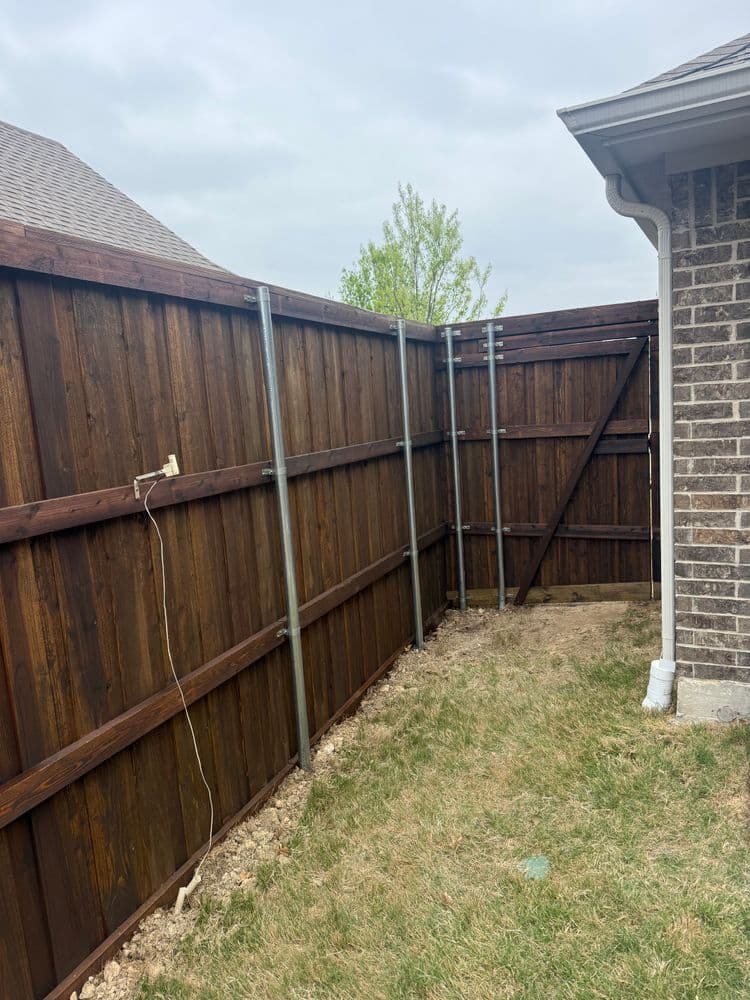 Wooden fence corner with metal support posts, grass, and a house wall in the background.