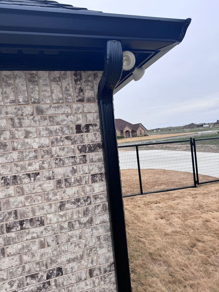 Black gutter and downspout on a brick house exterior with a fenced yard in the background.