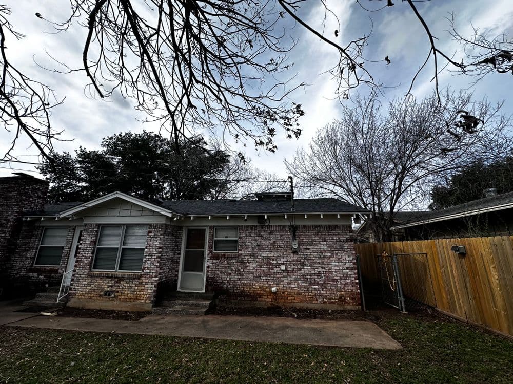 Red brick house with a dark roof, bare trees, and a wooden fence under cloudy skies.