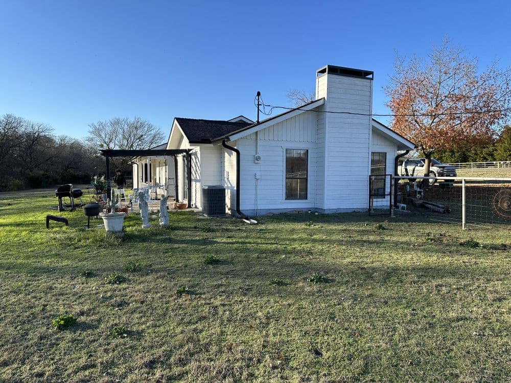 Charming white farmhouse with porch in a rural setting surrounded by green grass and trees.