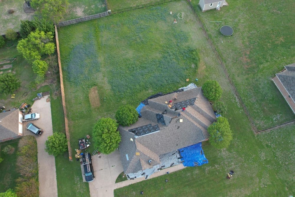 Aerial view of a house with a blue tarp on the roof and green lawn, surrounded by trees.