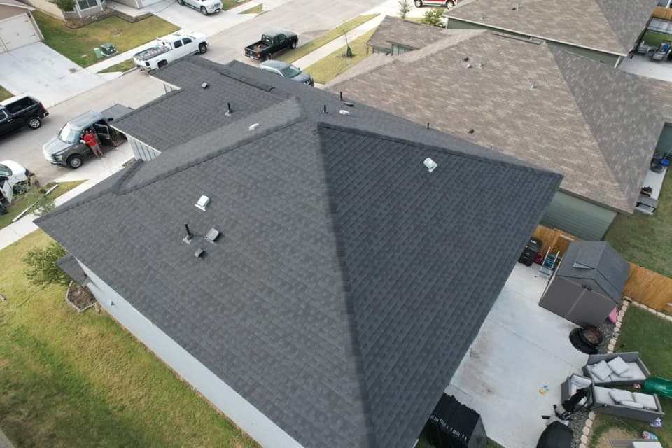 Aerial view of residential rooftops with dark shingles and nearby vehicles in a suburban area.