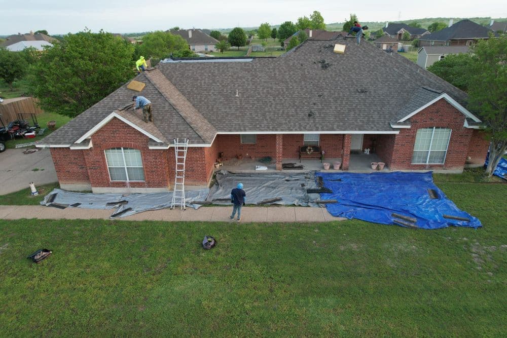 Roofers working on a home, using ladders and tarps, with a grassy yard and residential area visible.