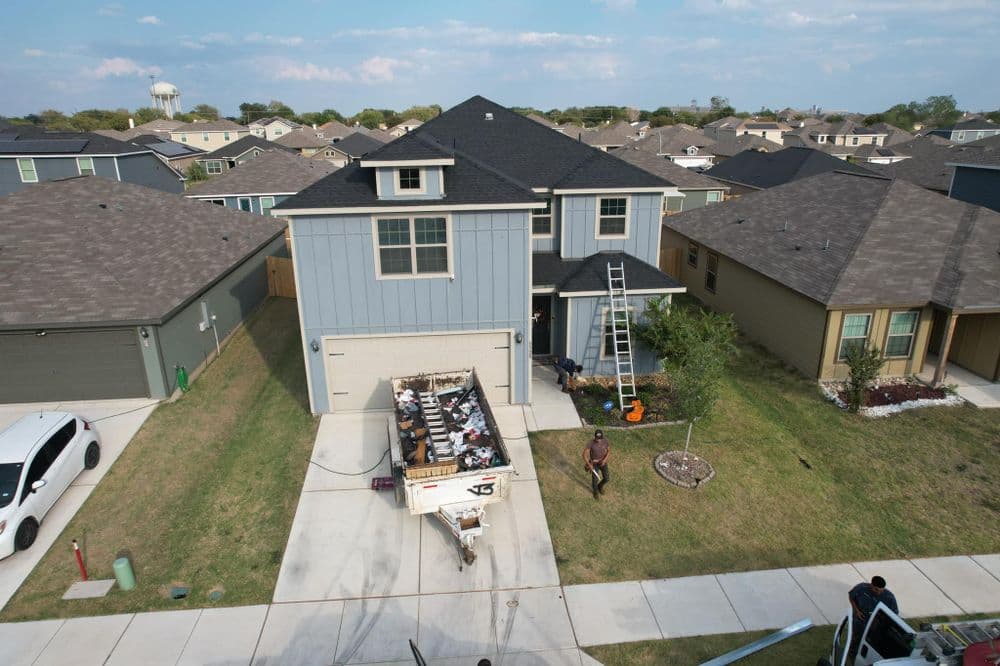 Two-story blue house with a ladder, driveway, and moving truck outside in a residential area.