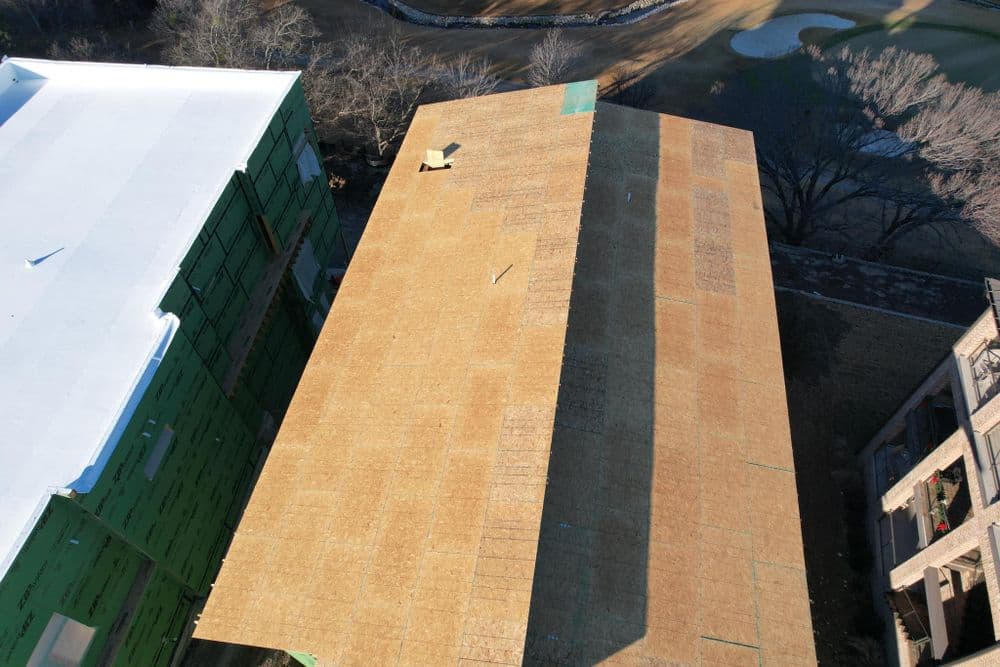 Aerial view of a building under construction, featuring plywood roofing and green insulation.