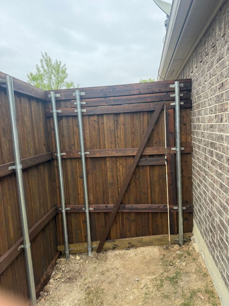 Wooden fence with metal supports, partially open entrance, set against a brick wall.