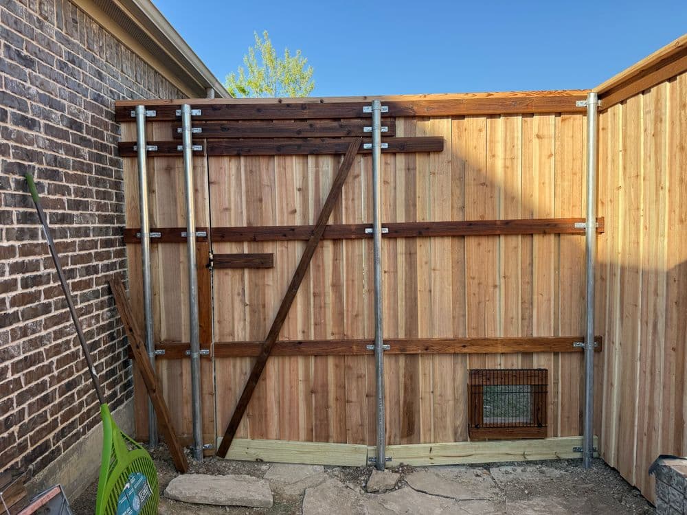 Wooden fence with metal supports and a small access gate, set against a brick wall.