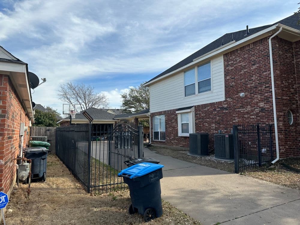 Residential driveway with a fence, basketball hoop, and trash bins beside a brick house.