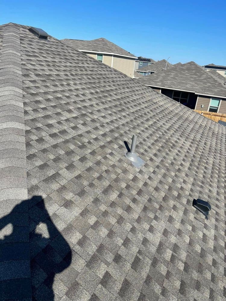 Asphalt shingle roof with vent pipes against a clear blue sky and suburban houses in background.