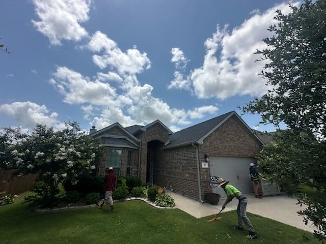 Home exterior with brick facade, green lawn, and workers maintaining landscaping under a cloudy sky.