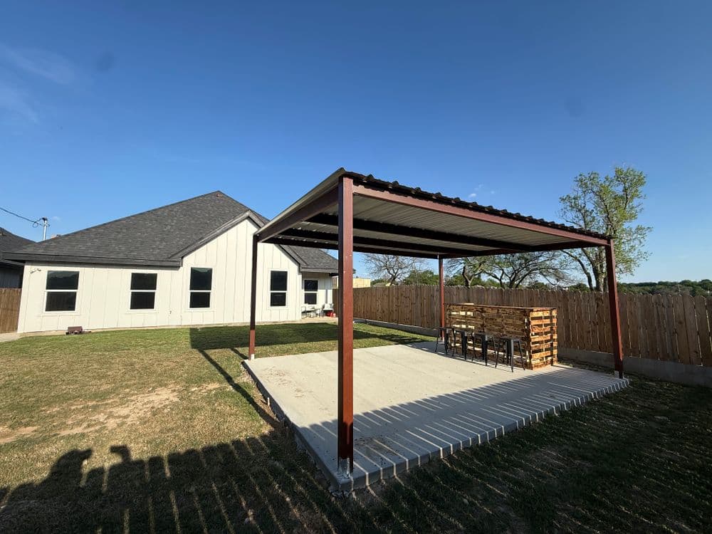 Modern backyard patio with metal pergola and seating area near a house. Clear blue sky.